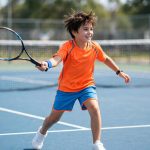 Young child playing tennis on a hard court wearing white tennis shoes.